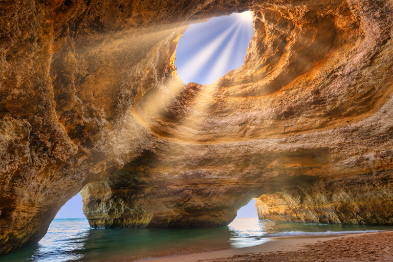 Vue sur le plafond de la grotte de Benagil avec rayons de soleil et mer en arrière plan.